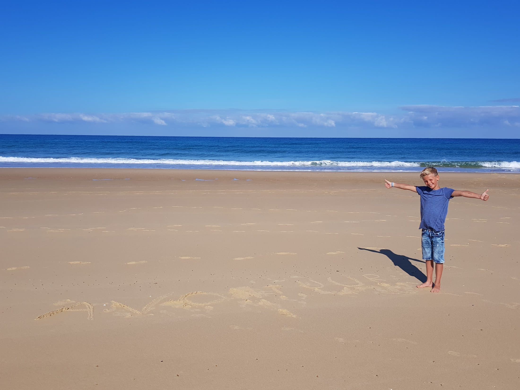 Vrolijke jongen wijdbeens met duimen omhoog op het strand