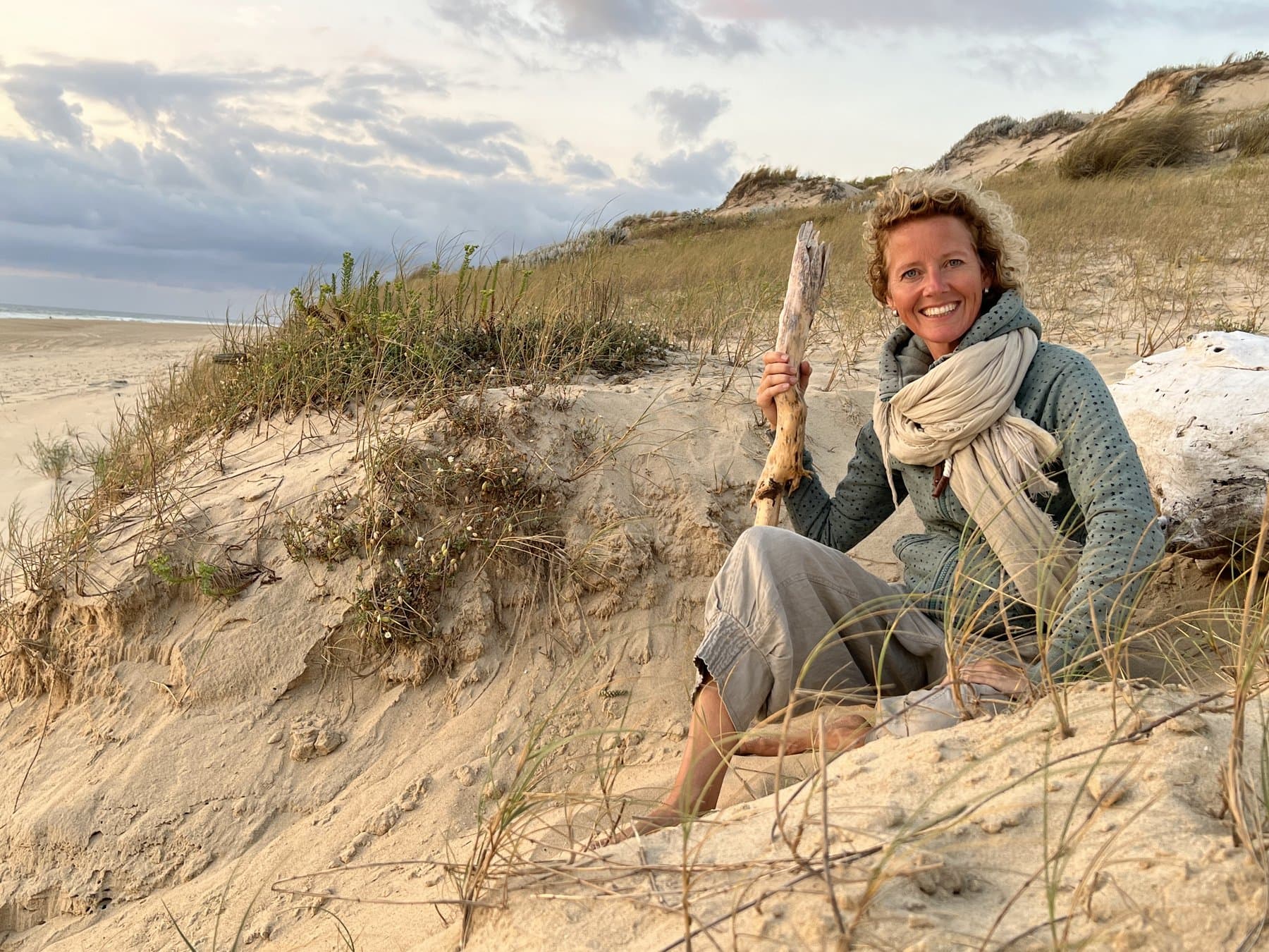 Simone zit lachend in de duinen met een stuk drijfhout in haar hand
